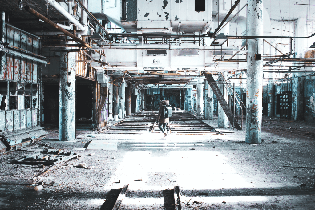 A man in a duffle coat and backpack is walking through a ruined factory